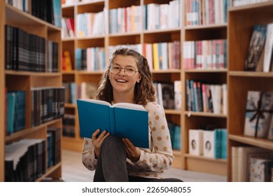 Teen girl among a pile of books. A young girl wearing glasses reads a book with shelves in the background. She is surrounded by stacks of books. Book day. - Powered by Shutterstock