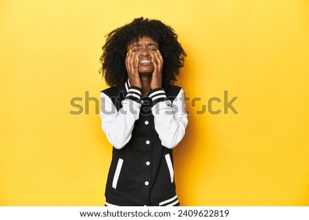Teen girl in baseball jacket, yellow studio background whining and crying disconsolately.