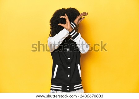 Teen girl in baseball jacket, yellow studio background keeping two arms crossed, denial concept.