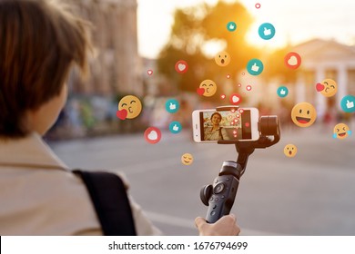 Teen girl blogger vlogger record vlog streaming video hold phone on selfie stick in urban city. Young female vlogger shoot social media blog on smartphone get likes emoji, over shoulder closeup view.