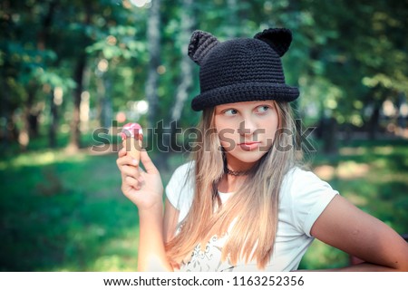 teen girl blond long hair in black crochet hat with cat ears in summer outdoors walking eating icecream smiling laughing green background