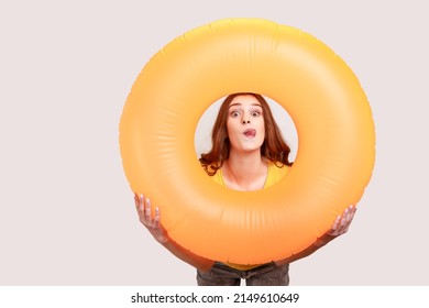 Teen Girl Of With Brown Wavy Hair In Yellow T-shirt Holding Orange Rubber Ring, Having Fun On Resort, Looking At Camera And Showing Tongue Out. Indoor Studio Shot Isolated On Gray Background.