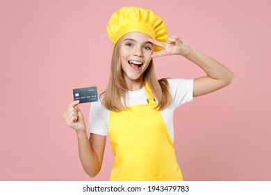 Teen girl chef cook confectioner housewife mother's helper baker in yellow apron white t-shirt cap hold credit bank card cover eye with victory gesture isolated on pink background Food cake concept
