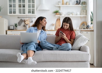 Teen girl child daughter and young mother using gadgets together, sitting on sofa at home. Curious parent mom peeping who kid chatting in social networks. Family and digital technologies concept - Powered by Shutterstock