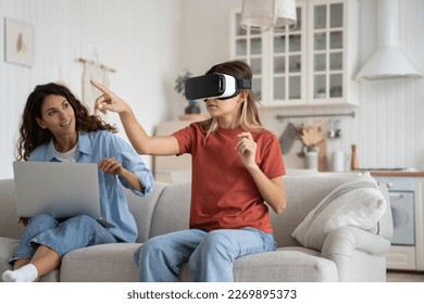 Teen girl child wearing virtual reality goggles at home, schoolgirl kid experiencing remote learning with VR while sitting with working remotely mother beside. Role of gadgets in modern family life - Powered by Shutterstock