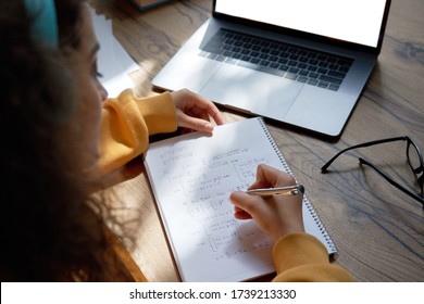 Teen Girl College Student Wear Headphones Studying From Home Writing In Workbook Solving Equations Learning Math Sits At Desk. Teenage School Pupil Learn Online On Laptop, Close Up Over Shoulder View.