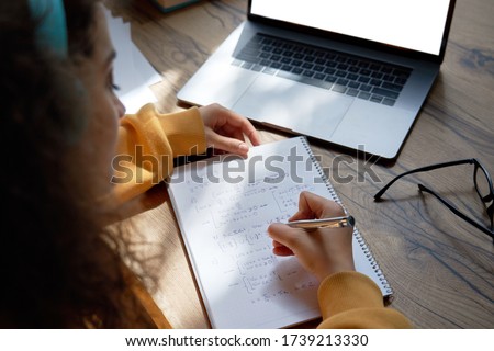 Teen girl college student wear headphones studying from home writing in workbook solving equations learning math sits at desk. Teenage school pupil learn online on laptop, close up over shoulder view.