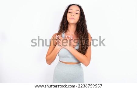 teen girl with curly hair wearing grey sport set over white background smiling with hands on chest with closed eyes and grateful gesture on face. Health concept.