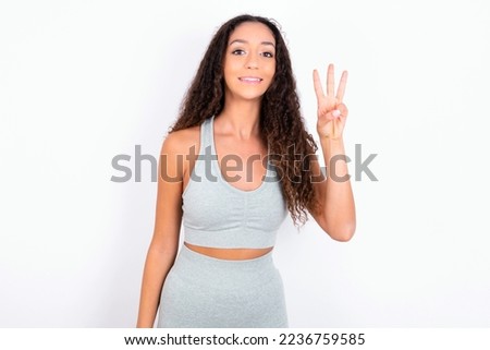 teen girl with curly hair wearing grey sport set over white background showing and pointing up with fingers number three while smiling confident and happy.