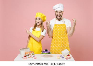 Teen Girl Dad Chef Cook Confectioner Baker Wear Yellow Apron Cap At Table Point Finger Aside On Workspace Do Winner Gesture Isolated On Pink Background. Mousse Cake Food Workshop Master Class Process