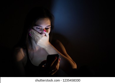 Teen girl excessively sitting at the phone at home. She is a victim of online social networks. Sad teen checking phone sitting on the floor in the living room at home with a dark background.