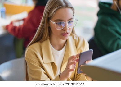 Teen girl gen z student using mobile phone looking at smartphone sitting at desk in university college campus classroom. Young blonde woman holding cellphone modern tech in university. - Powered by Shutterstock