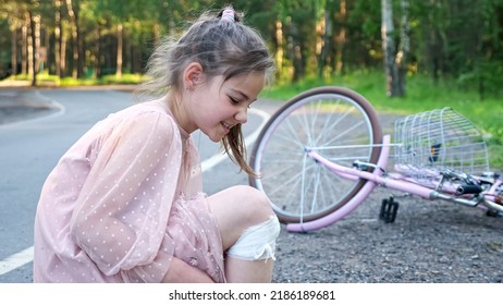 Teen Girl Grimaces Because Of Pain In Leg. Girl Wearing Pink Dress Falls From Pink Bicycle Trying To Learn To Ride Bike. Schoolgirl Has Injury On Knee Closeup