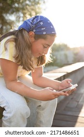 Teen girl holding a mobile phone while sitting on a bench outdoors. Young woman responds to sms, chatting. Stylish image, blue bandana. Soft focus. concept of online, follower, social media. Sunset.