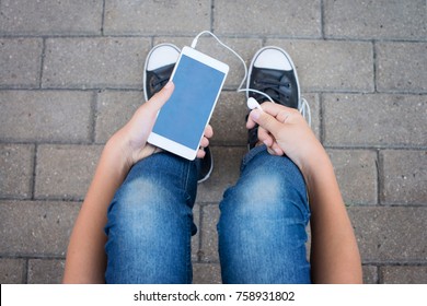 Teen girl in jeans and sneakers sitting outdoors, using a smartphone in the hands, close up, white smart phone.
