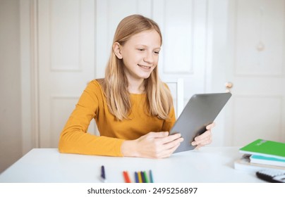 A teen girl with long blonde hair sits at a white desk in a home office, using a tablet computer. She is smiling and looking at the screen. A stack of books and some colored pencils are on the desk. - Powered by Shutterstock