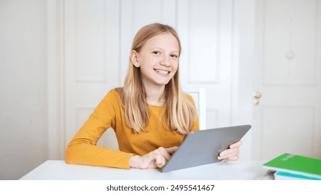 A teen girl with long blonde hair sits at a table, smiling as she uses a tablet computer. The table is white, and the background is a white wall with a closed door. - Powered by Shutterstock