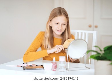 A teen girl with long blonde hair sits at a vanity table, brushing her hair in front of a mirror. She is wearing a mustard yellow long-sleeved shirt and has a relaxed and happy expression on her face. - Powered by Shutterstock