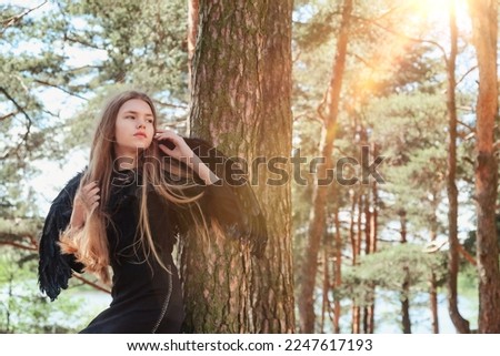 Teen girl with long hair in black angel wings posing in fairy tale forest, looking away. Young pretty lady at mysterious woodland. Vintage retro image, fantasy concept. Copy space for advertising text