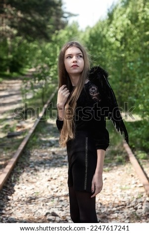 Teen girl with long hair in black angel wings posing in mysterious forest, looking up. Young pretty lady at fairy tale woodland. Vintage retro image, fantasy concept. Copy space for advertising text