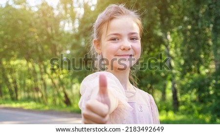 Teen girl looks straight on big forest pathway. Schoolgirl wearing pink dress shows thumb-up smiling closeup. Girl enjoys walking