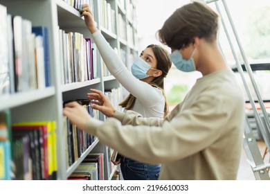 Teen Girl In Medical Mask Choosing Books In Library Near Friend On Blurred Foreground