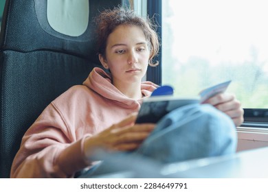 teen girl in pink hoodie sits by window in train car, reads magazine, thoughtfully. Traveling on student vacation. teen girl in train reading book and using mobile phone - Powered by Shutterstock