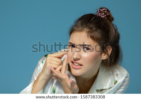 Teen girl with problem skin look at pimple, blue background. Morning theme, skin care. Teenager with acne
