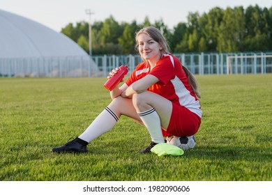 Teen girl in red football strip sitting on soccer ball holding bottle of water at sunset, smiling to camera
