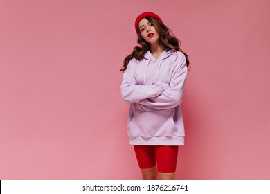 Teen Girl In Red Hat And Cycling Shorts Poses On Pink Background. Portrait Of Curly Attractive Woman With Bright Lips Dressed In Purple Oversized Hoodie On Isolated.