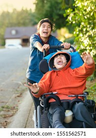 Teen Girl Running And Smiling While Pushing Disabled Little Boy In Wheelchair Outdoors 