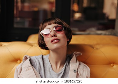 Teen girl with short hair posing on couch inside. Charming brunette woman in pink sunglasses and denim beige outfit looking into camera in cafe..