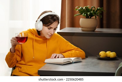 Teen girl is sitting at the table, reading book and listening music while drinking tea on kitchen. Headphones on her head. Wellness rest and relaxation hobby - Powered by Shutterstock