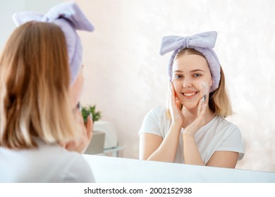 Teen Girl Washing Face Portrait Reflection In Mirror In Morning Bathroom. Young Blonde Woman Applies Facial Cleaner For Skin Wash. Self Care Morning Bathroom Routine