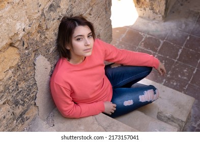 Teen Girl Wearing Pink Long Sleeve Shirt Looking Into A Camera. Mockup. Serious Teenager Girl Sitting Outdoor On A Stairs Spending Her Time Alone. Adolescence Concept