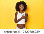 Teen girl in white tank top, yellow studio background has friendly expression, pressing palm to chest. Love concept.