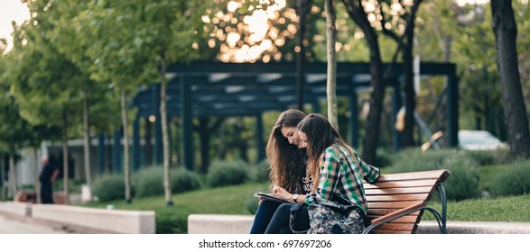 Teen Girls First Day Of School Outfit.Showing The Old Funny Pictures On Tablet.Evergreen Memories.Friendship Goals.Really Happy To See Those Old Pictures.Sitting On Wooden Bench In Park.Back To School