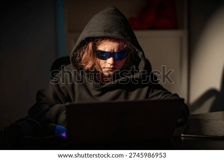 Teen hacker working on computer in dark room. Kid hacker using laptop in a dark room. Teenage hacker working online in secret. Child hacker cyber security theme.