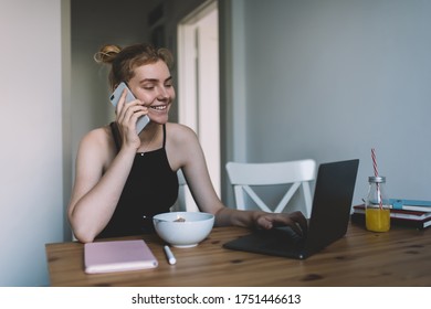 Teen Happy Female With Blond Hair Talking On Mobile Phone And Using Laptop While Sitting At Wooden Table In Tidy Room