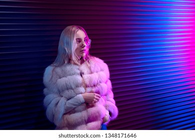 Teen hipster girl in stylish glasses and fur standing on purple street neon light wall background, female teenager fashion model pretty young woman looking at night club city light glow, back to 80s - Powered by Shutterstock