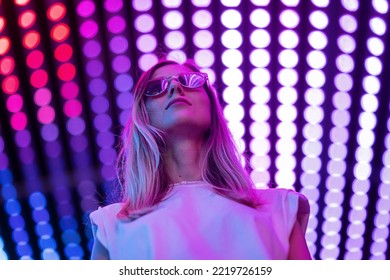 Teen hipster girl in stylish glasses standing on purple tunnel with neon light wall background, female teenager fashion model pretty young woman looking at night club city light glow