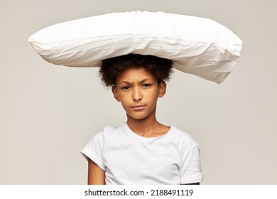 Teen Kid Boy With Afro Hair Suffering From Insomnia Looking At Camera With Upset Annoyed Face Expression In White Mockup T-shirt With Copy Space For Your Text On Gray Background With Pillow On Head