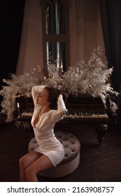 teen leisure. young brunette beautiful girl in white glamour dress is sitting sensual near black grand piano with dried flowers on vintage room background with mirror. lifestyle concept, free space