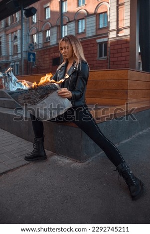 teen rebel walk leisure. young girl in black leather jacket with burning newspaper in hands is sitting fashion on the street glass cafe background and looking straight. lifestyle concept, free space