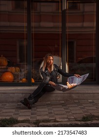 Teen Rebel Walk. Young Blonde Girl In Black Leather Jacket With Burning Newspaper In Hands Is Sitting Fashion With Long Legs On Street Glass Cafe Background And Smiling. Lifestyle Concept, Free Space