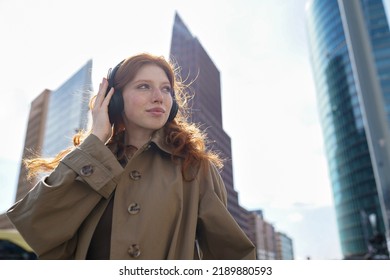 Teen Redhead Girl Standing On Urban Street Skyscrapers Background Wearing Headphones Listening To Music, Audio Book, Travel Guide Or Podcast While Walking In Big City.