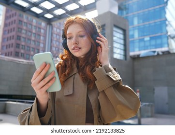 Teen Redhead Girl Standing On Urban Street Skyscrapers Background Wearing Headphones Using Smartphone Listening To Music, Audio Book, Travel Guide Or Podcast While Walking In Big City.