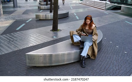 Teen redhead hipster girl student wearing headphones using laptop and smartphone in the city online learning outdoors, elearning outside watching webinar sitting in urban street. - Powered by Shutterstock