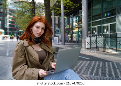 Teen redhead hipster girl student using laptop computer modern technology device on city street searching information, online learning outdoors, elearning outside sitting in urban park.