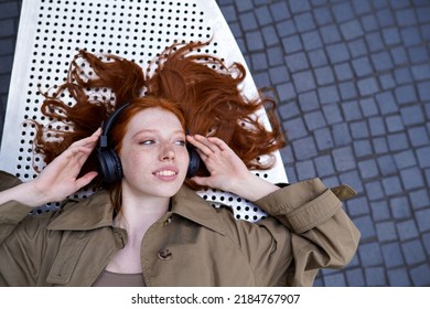 Teen Redhead Hipster Girl Wearing Headphones Listening To Mobile Streaming Music Mix, Audio Book, Travel Guide Or Podcast Enjoying Modern Sound Tech Outdoors Looking At Copy Space. Top View